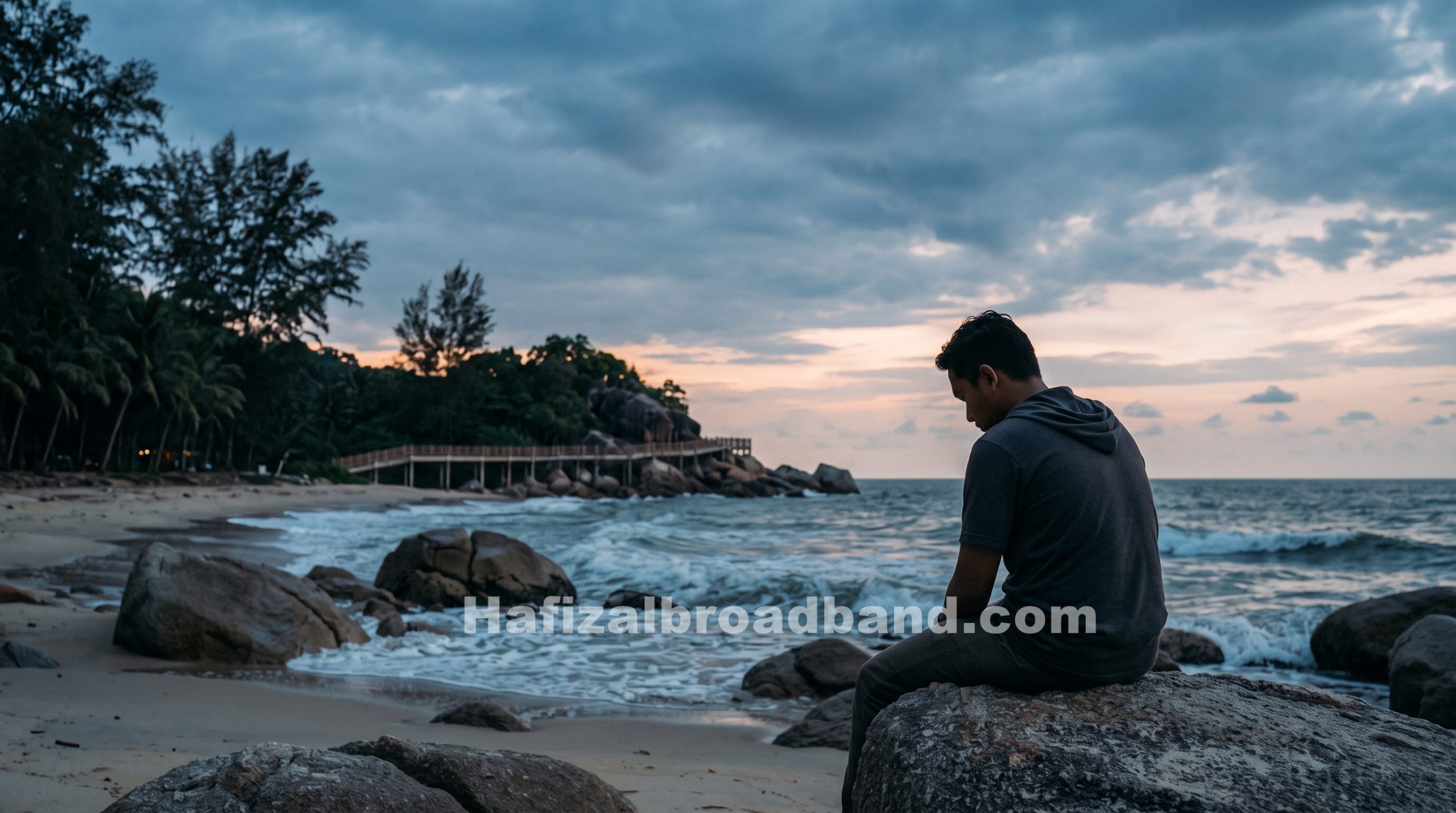 Seorang lelaki duduk termenung di atas batuan Pantai Teluk Cempedak Kuantan ketika waktu senja melambangkan refleksi jiwa dan penerimaan takdir.